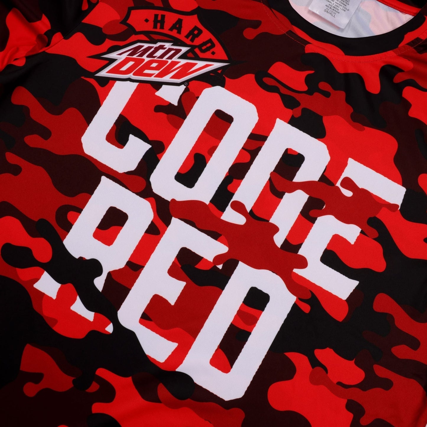 A close-up view of the chest area of a red and black camouflage short-sleeved t-shirt. The 'Hard Mtn Dew' logo is visible in the upper left, with the large white block letters 'CODE RED' printed prominently below it, in two lines. The textured fabric and printed camouflage pattern are clear.