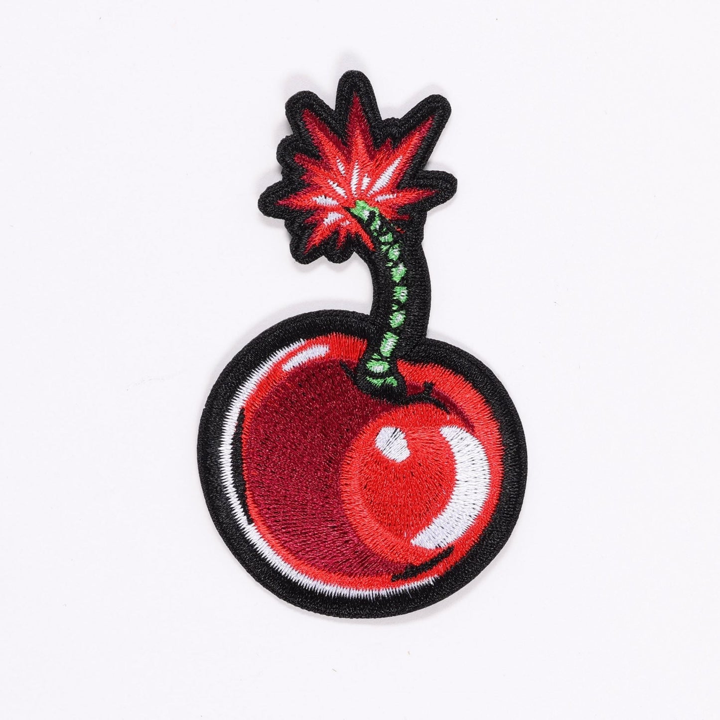 An embroidered patch depicting a "cherry bomb." It features a round, red bomb with a black outline and a stylized fuse that resembles a green cherry stem with black dots and red, starburst-shaped flames outlined in black at the top. A white crescent shape suggests a highlight on the bomb's surface.