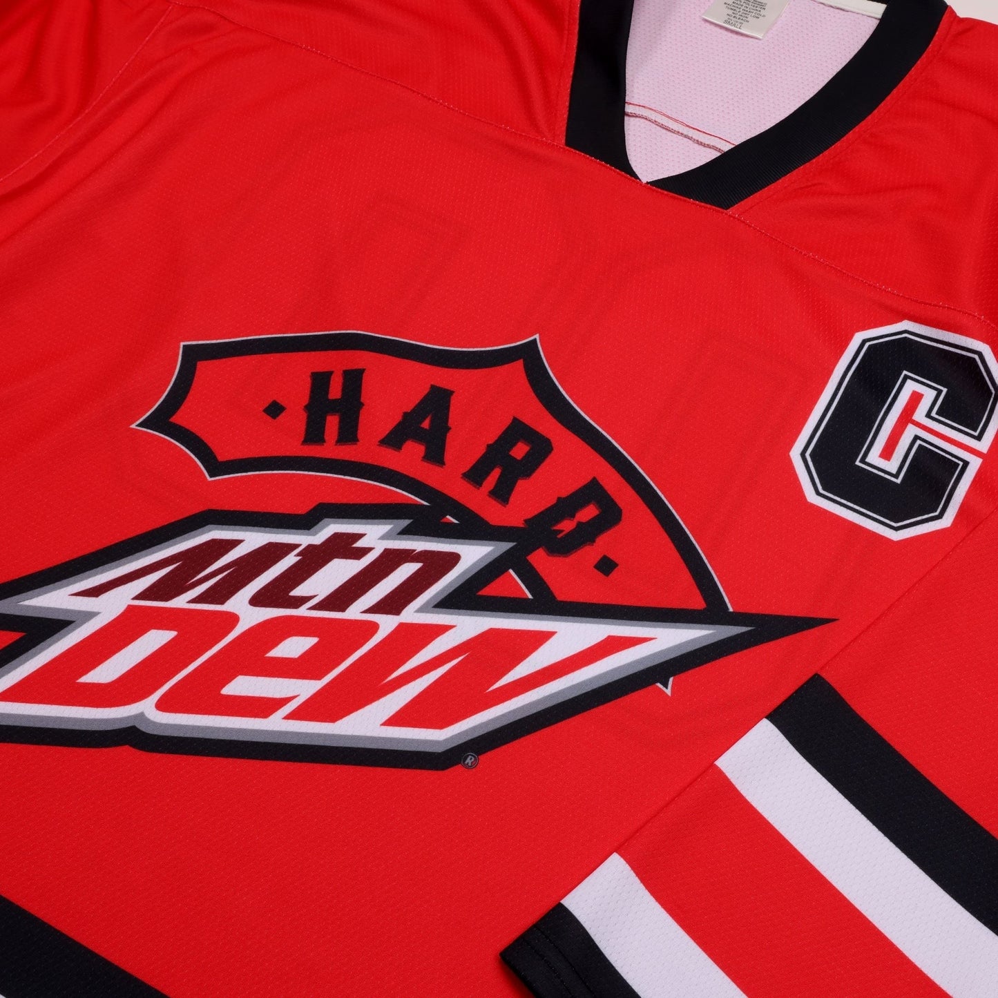 A close-up shot of the front of a red hockey jersey, focusing on the chest area. The 'Hard Mtn Dew' logo is prominently displayed, with 'Hard' in white on a red banner above the white, red, and black Mtn Dew logo. To the right, a large black 'C' with a white outline is visible. The jersey has a black V-neck collar and visible stitching, suggesting fabric texture.