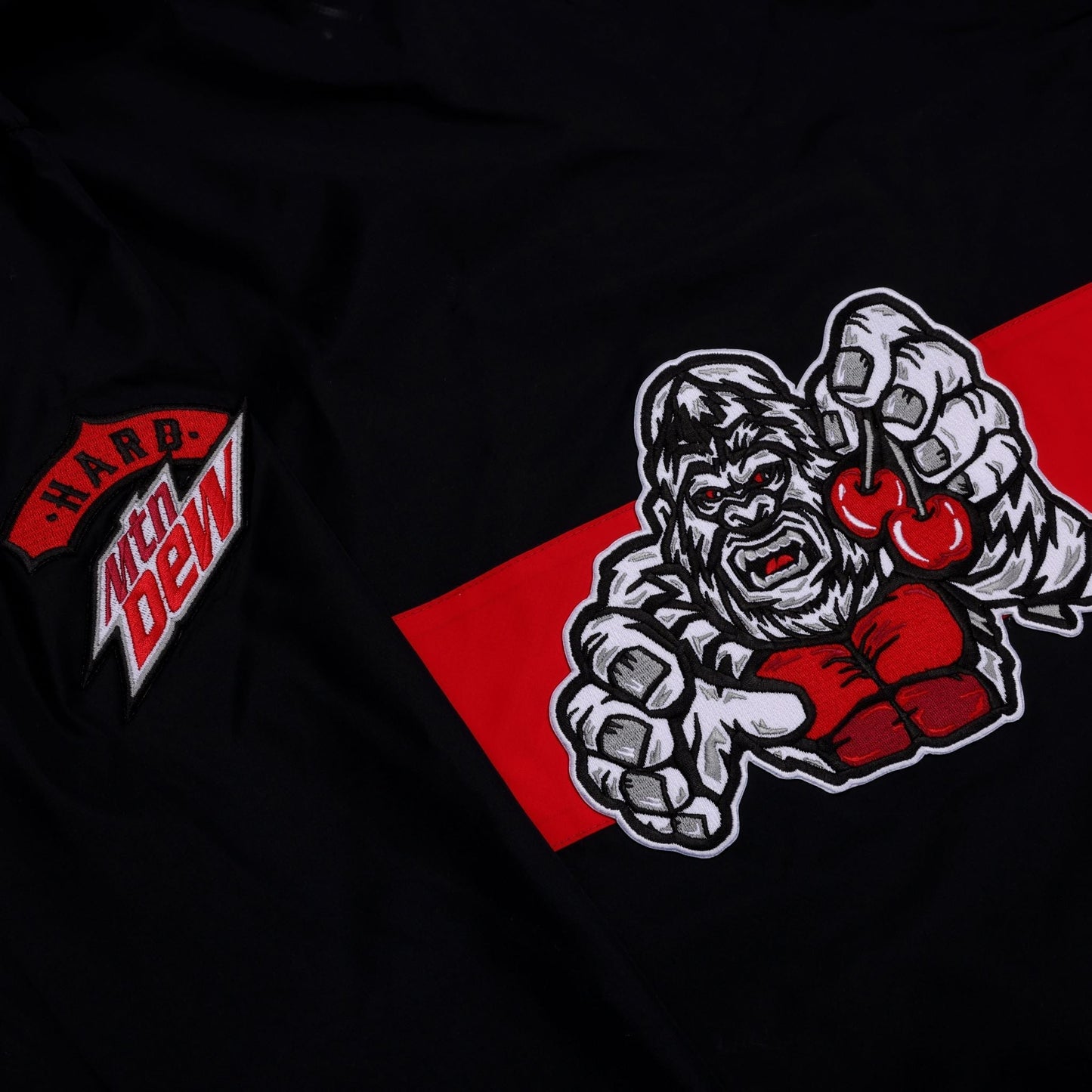 A close-up view of a black windbreaker, focusing on the left chest and shoulder area. The 'Hard Mtn Dew' logo is visible as an embroidered patch on the left side. A red diagonal stripe extends from the lower right of the frame, upon which a graphic of an aggressive gorilla with red eyes holding two red cherries in its right hand.