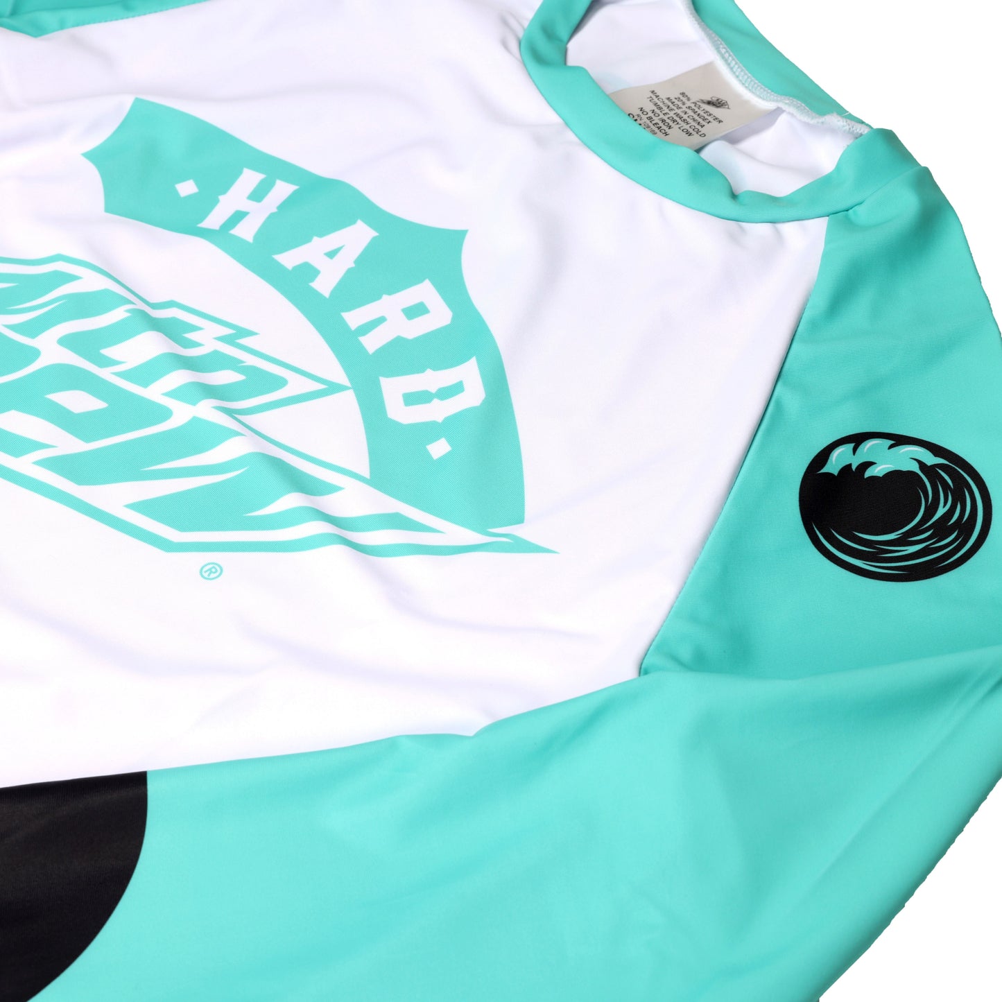 Stylized angle view of the long sleeve rash guard laying flat, showing the top portion of the right side. The front features a white body with a large teal Hard Dew logo on the chest. The raglan-style shoulder is teal blue with a black circular wave desgin.