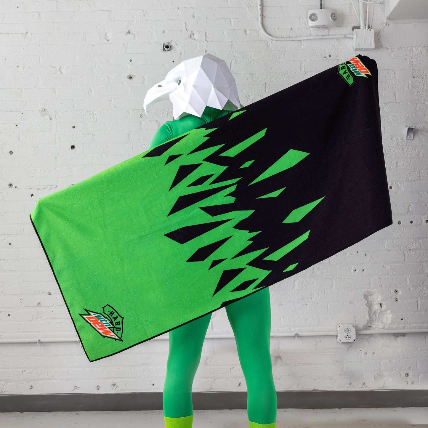 Model wearing an eagle head mask holds up a Hard Mountain Dew towel, demonstrating that the towel is approximately the length of the mask's wingspan. The towel's design and logo are clearly visible.