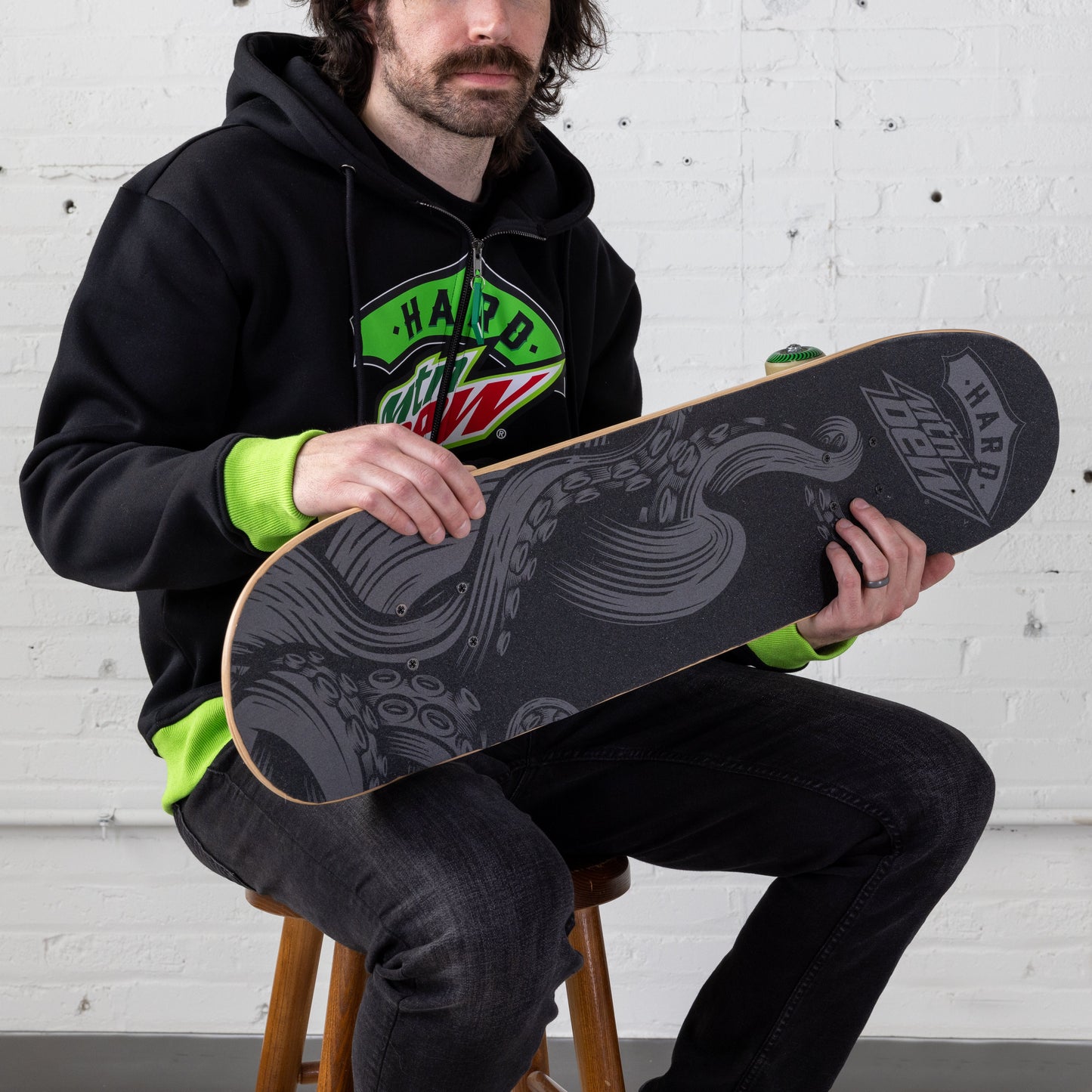 Model holding a skateboard with black and grey grip tape featuring an octopus tentacle arm design and the Hard Mountain Dew logo.