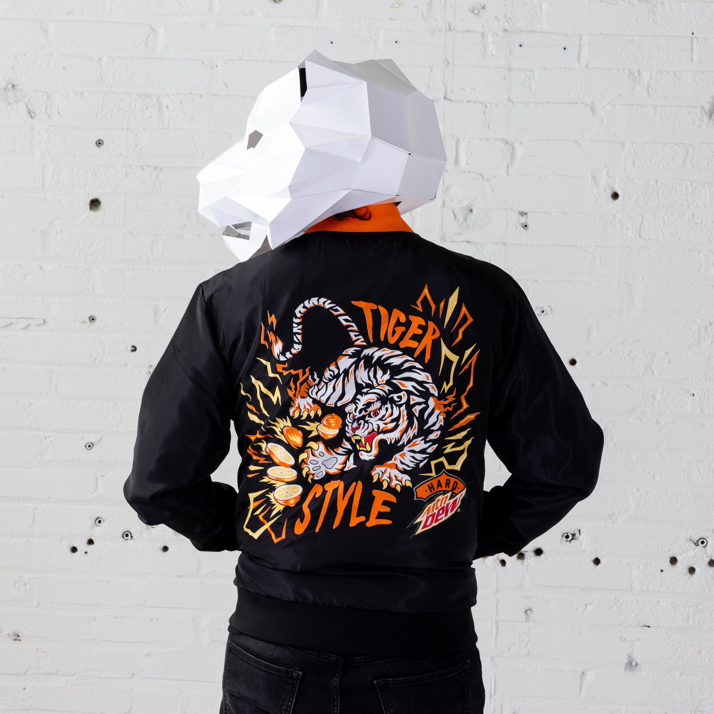 Model wearing a Hard Mountain Dew LiveWire bomber jacket with a white tiger mascot-like mask over their head. The back of the jacket features vibrant colors and a distinctive design with a white tiger.