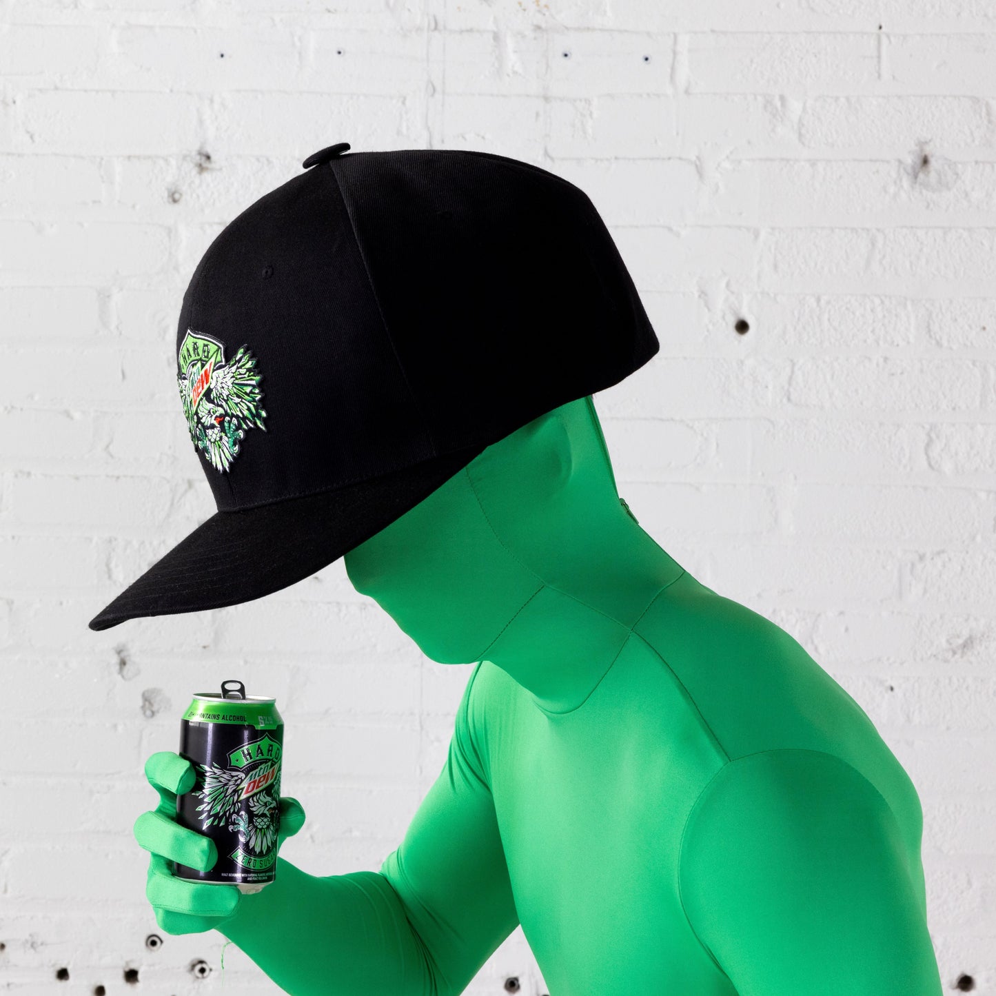 Profile view of a model in a full face-covering bright green skin suit, wearing the oversized black baseball hat and holding a Hard Mountain Dew can. The hat features neon green, white, and red embroidery of an eagle and the Hard Mountain Dew logo.