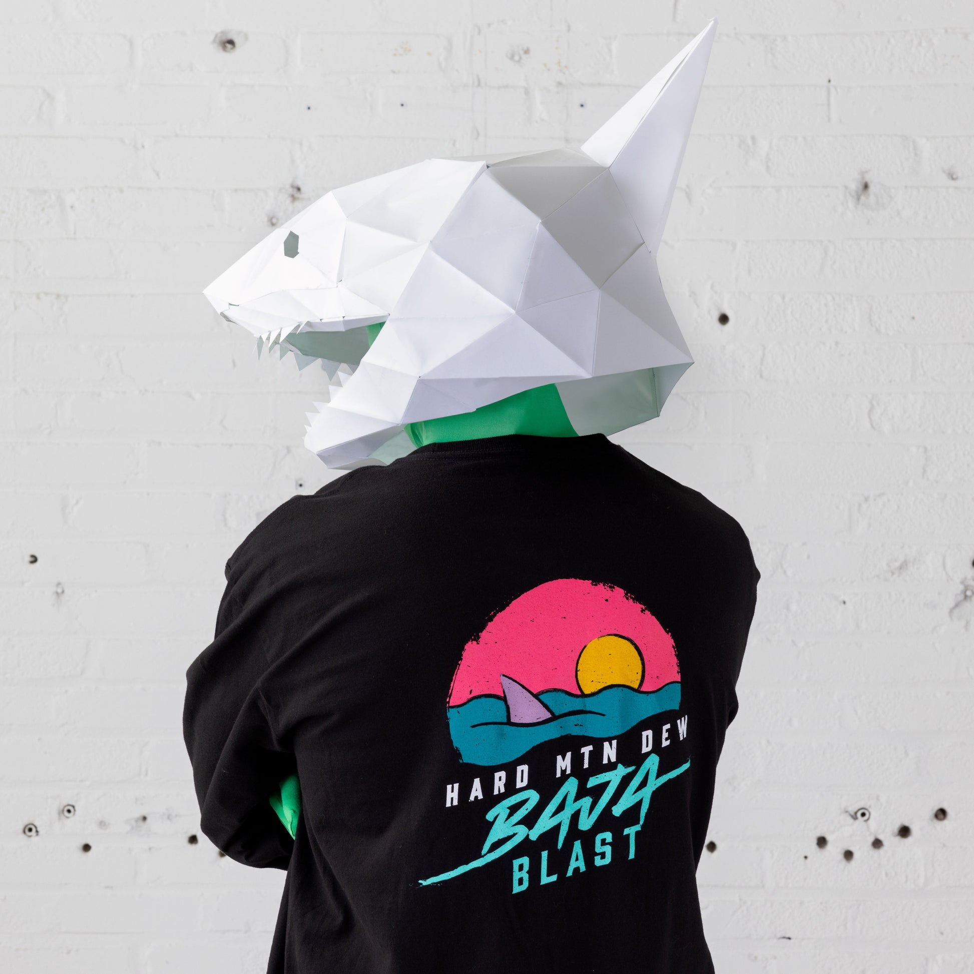 Back shot of the top half of a black long sleeve tee worn by a model with a shark mask. The shirt features a large retro 90s design with a shark fin emerging from teal water, a pink horizon, and an orange sun within a circular shape. The text 'Hard Mountain Dew' and 'Baja Blast' in teal blue is positioned below the illustration.
