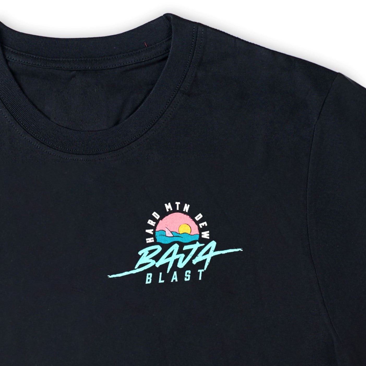 Close-up of the front artwork on the black Baja Blast t-shirt. The design features a retro 90s graphic of a shark fin emerging from teal water with a pink horizon and orange sun, all within a circular shape. The Hard Mountain Dew text arches around the top of the circle, and Baja Blast is written underneath.