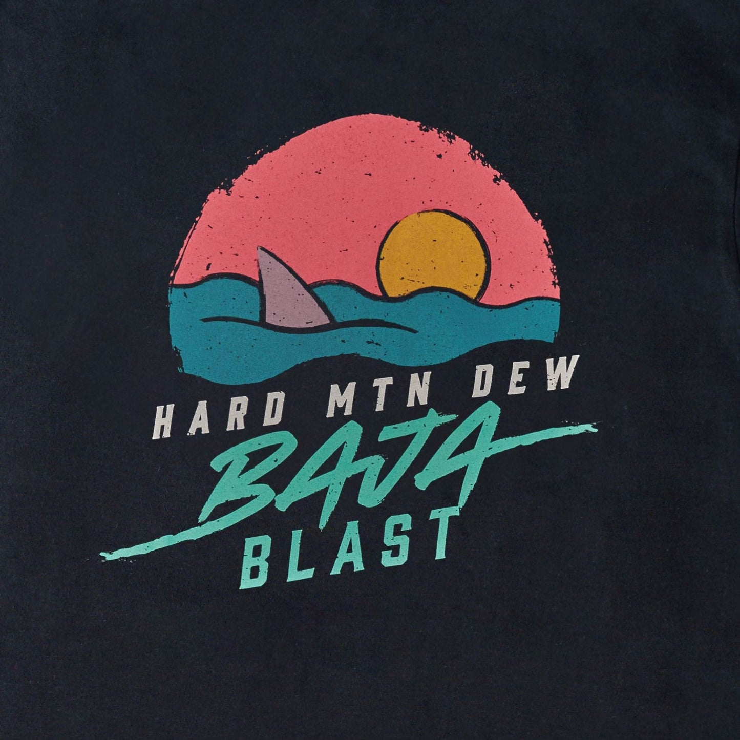 Close-up of the back art detail on the black long sleeve tee. The design features a shark fin emerging from teal water, with a pink horizon and orange sun within a circular shape. The text 'Hard Mountain Dew' and 'Baja Blast' in teal blue is positioned below the illustration, showcasing the retro 90s aesthetic.