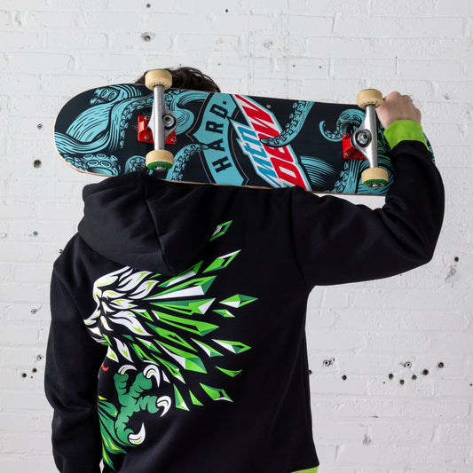 Model holding a skate deck over their shoulder. The deck features a teal, black, and red Hard Mountain Dew logo surrounded by octopus tentacle arms. The wheels of the skateboard are off-white with green and black details on the sides. The model is wearing a Hard Mountain Dew zip-up sweatshirt with a large eagle design on it. Note: Wheels not included.