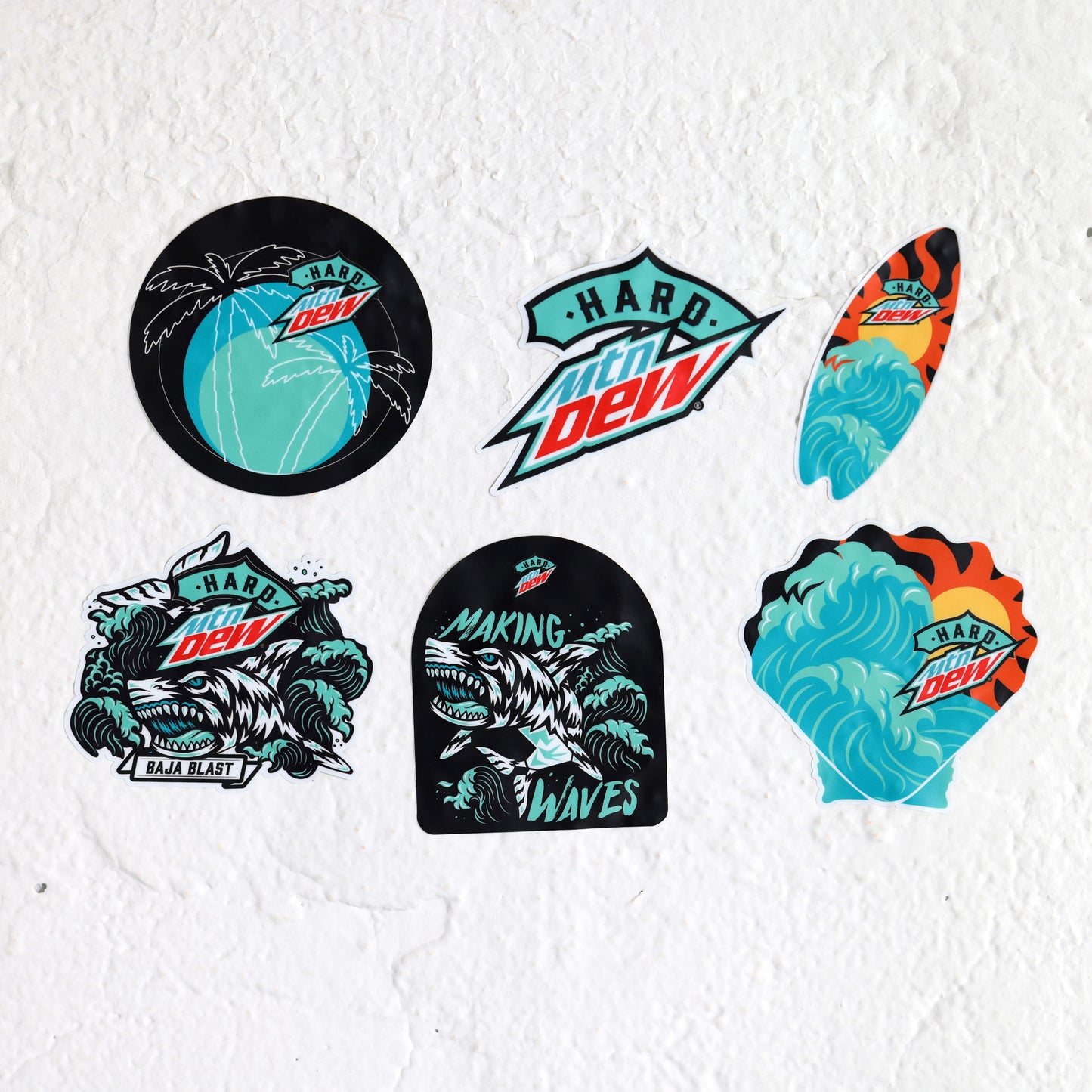 Pack of seven stickers featuring Baja Blast-inspired designs. Includes: a circular sticker with palm trees and a small logo; a boogie board-shaped sticker with a teal wave and a yellow and orange sun; a shark Baja Blast mascot surrounded by waves; another shark sticker in a semi-circle shape; a wave and sun design in a clam shell shape; and a sticker with the Hard Mountain Dew logo in the Baja Blast teal color palette.