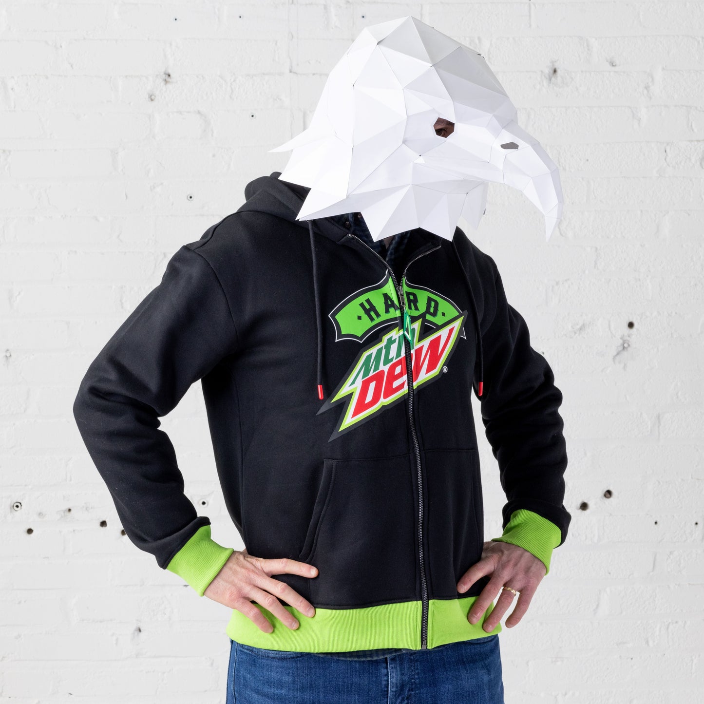Front view of a model wearing the Hard Mountain Dew zip-up hoodie sweatshirt. The hoodie features neon green knit cuffs and hem, red plastic tips on the drawstrings, and a custom neon green zipper pull. A large Hard Mountain Dew logo spans across the zipper on the chest. The model is also wearing a mascot-style full eagle mask on their head.
