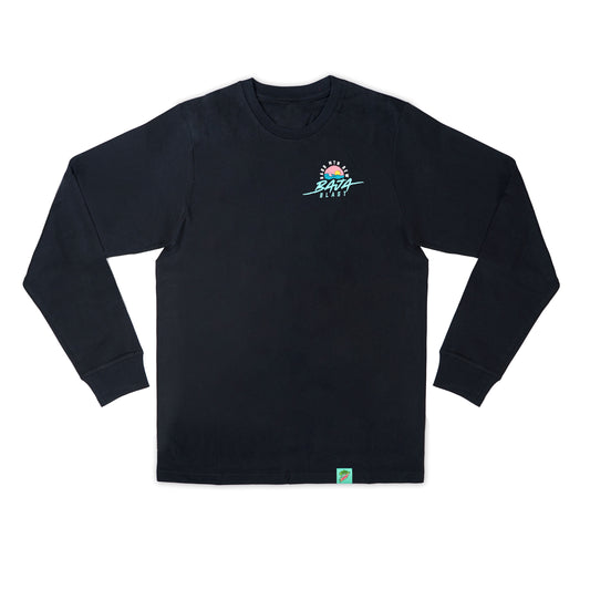 Flat lay of a black long sleeve tee featuring a retro 90s design. The artwork, positioned small on the left chest, shows a shark fin emerging from teal water with a pink horizon and orange sun, all within a circular shape. The Hard Mountain Dew text arches around the top of the circle, with Baja Blast written underneath. The bottom hem on the left side includes a small teal woven brand tag.