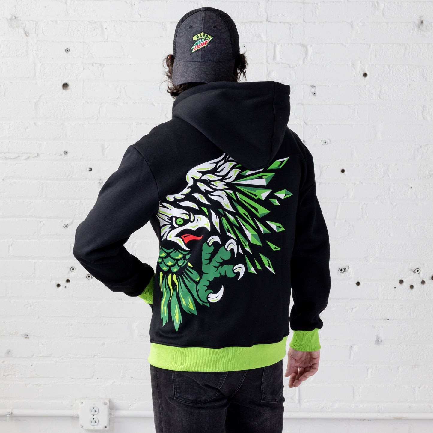 Back view of a model wearing the Hard Mountain Dew zip-up hoodie sweatshirt and a Hard Mountain Dew trucker hat. The hoodie features an oversized design of half an eagle head, body, claw, and wing in white, dark green, light green, and black.