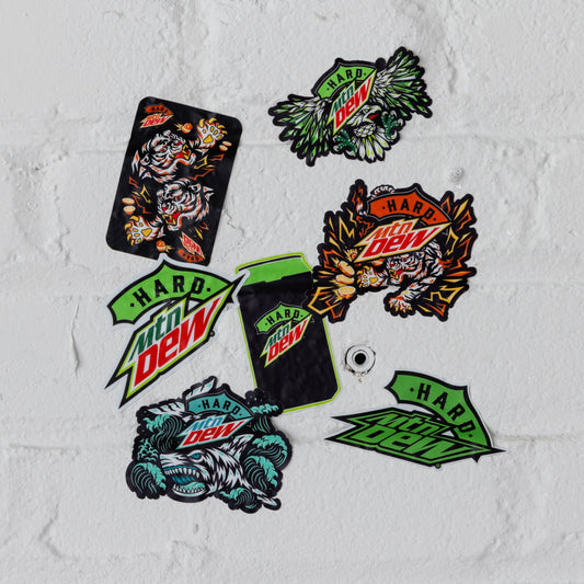 Pack of 7 Hard Mountain Dew stickers featuring various designs: the Baja Blast shark, the LiveWire tiger, the original eagle, a can shape, a full-color logo, and a stylized green and black logo. Each sticker showcases distinct artwork from Hard Mountain Dew cans.