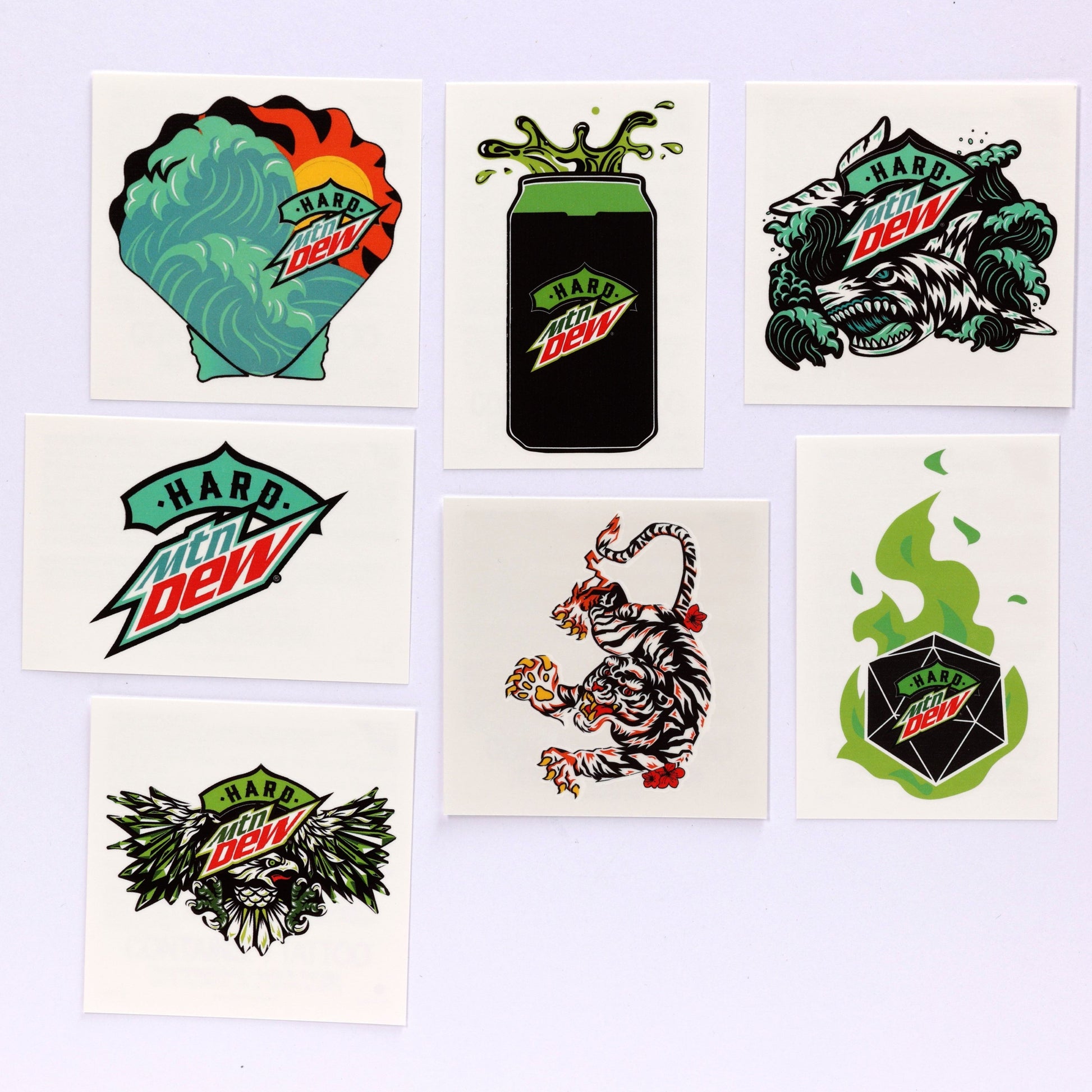 Seven temporary tattoos featuring Hard Mountain Dew branding: 1. Clamshell shape with teal wave and yellow/orange sun, Hard Dew logo over the artwork. 2. Hard Dew can with liquid splashing out. 3. Baja Blast shark in teal and black waves with Hard Dew logo. 4. Hard Mountain Dew teal Baja Blast logo. 5. Red, white, black, and orange LiveWire tiger. 6. Hexagonal black shape with Hard Dew logo and green flames. 7. Hard Mountain Dew eagle illustration with logo.