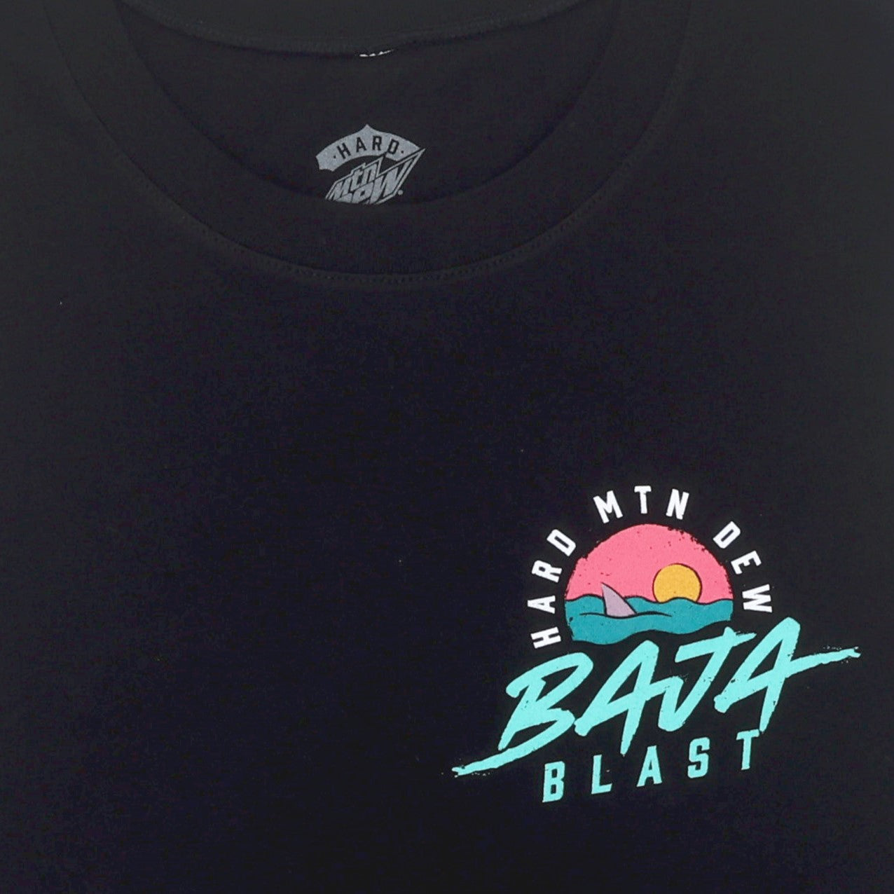Tight close-up of the front left chest artwork on a black muscle tank. The design features a small retro 90s graphic of a shark fin emerging from teal waves, with a pink sunset and orange sun in a circular shape. The Hard Mountain Dew Baja Blast text is integrated into the artwork.