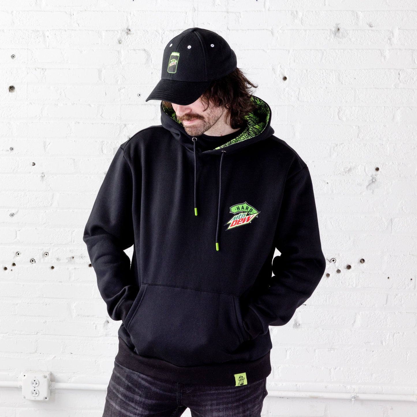 Front view of a model wearing a black Hard Mountain Dew hoodie with his hands tucked into the front kangaroo pocket. The model is also wearing a black dad hat.