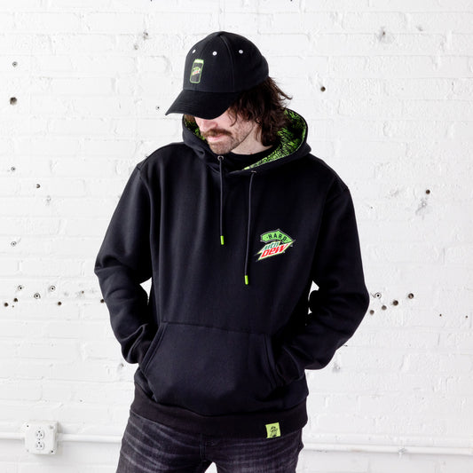 Front view of a model wearing a black Hard Mountain Dew hoodie with his hands tucked into the front kangaroo pocket. The model is also wearing a black dad hat.