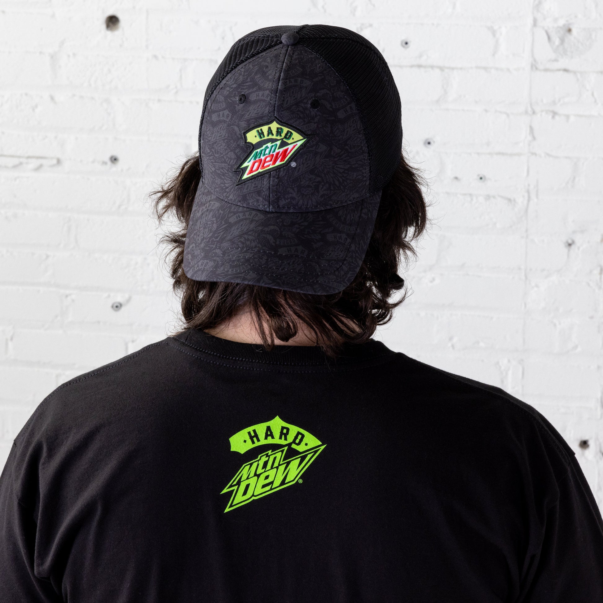 Model wearing a black trucker hat backwards, with the back of the hat and the model’s head visible. The hat features a tonal Hard Mountain Dew logo pattern printed across the front and on the bill, with the logo embroidered over the pattern.